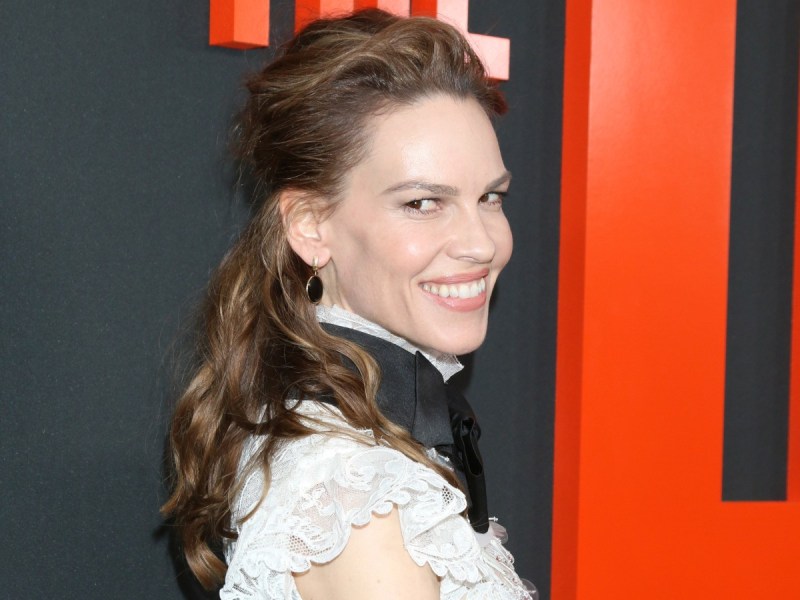 Hilary Swank smiles over her shoulder in white top with black collar