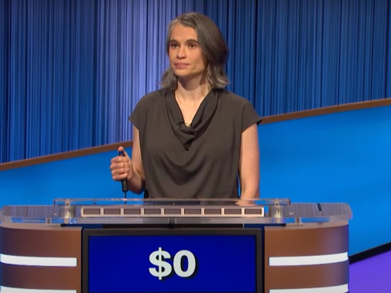 "Jeopardy!" contestant holds a buzzer in her hand at the podium. She is wearing a gray shirt