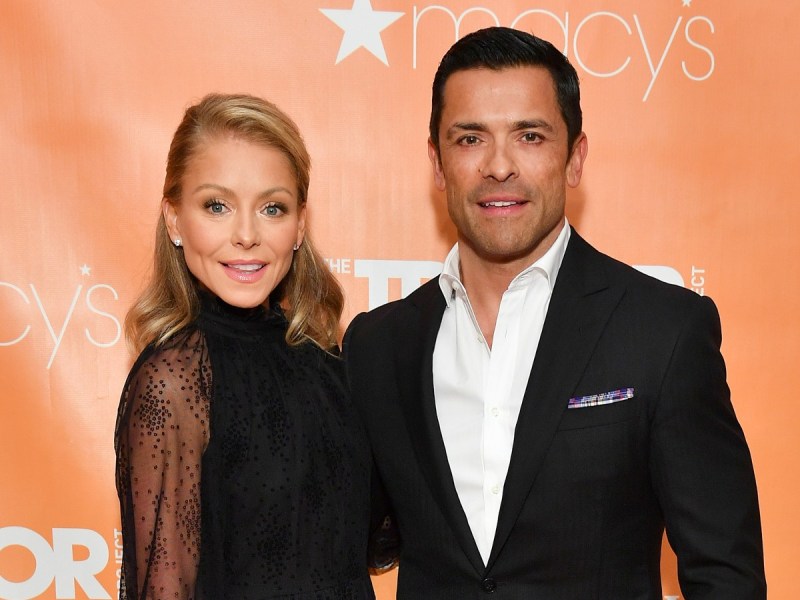Kelly Ripa (L) and Mark Consuelos smiling in black outfits against orange backdrop