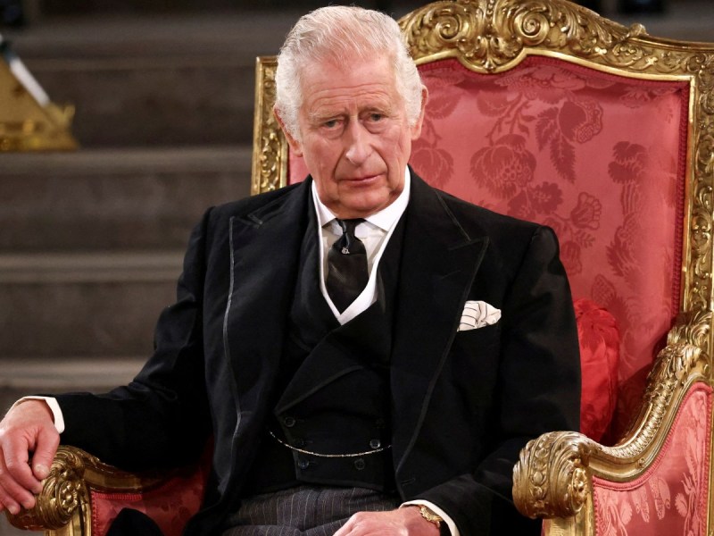 King Charles sits in red chair wearing black suit and tie