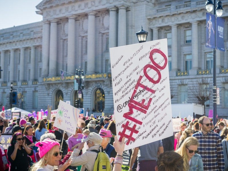 A white sign with the words "#METOO is being held up from a crowd of people in front of a U.S. government building