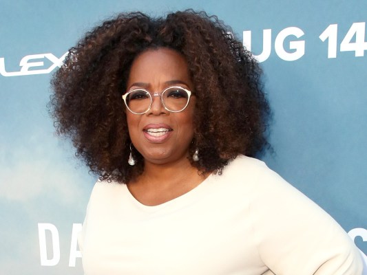 Oprah Winfrey smiles in white top against sky blue backdrop