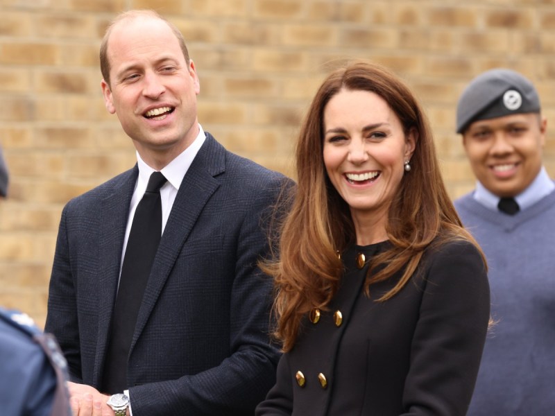 Prince William (L) in black suit laughing next to Kate Middleton, who is also wearing black and laughing