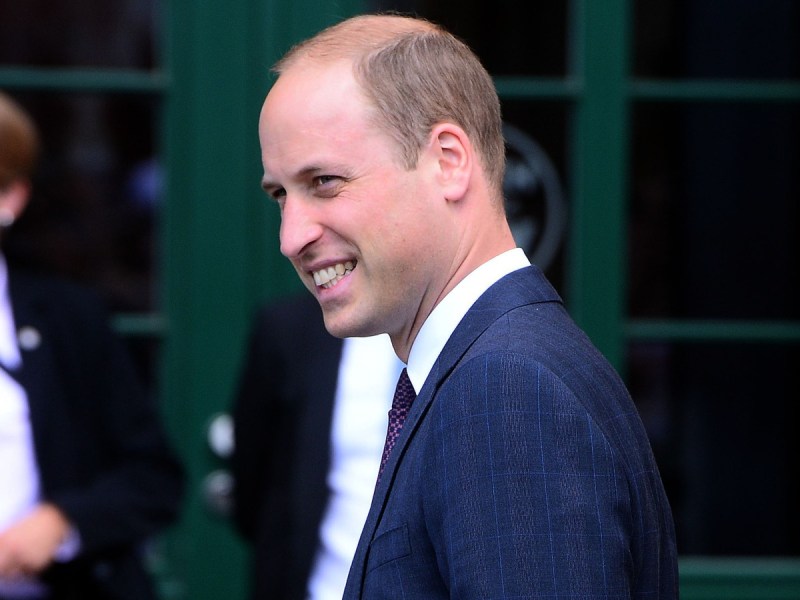 Prince William smiles in profile while wearing a navy blue suit against a green door