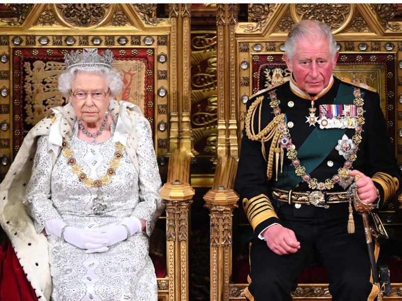 Queen Elizabeth (L) dressed in white on the throne seated next to then-Prince Charles, who is dressed in black with a green sash