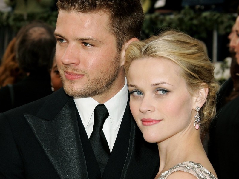 Closeup image of Reese Witherspoon (R) smiling next to Ryan Phillippe, who is wearing a black suit and tie
