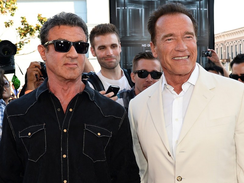Sylvester Stallone (L) in black top and sunglasses standing next to Arnold Schwarzenegger, who is wearing a cream-colored blazer