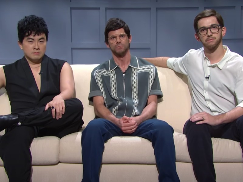 Three men sit on a couch on the 'SNL' stage. They all look intensely at the camera