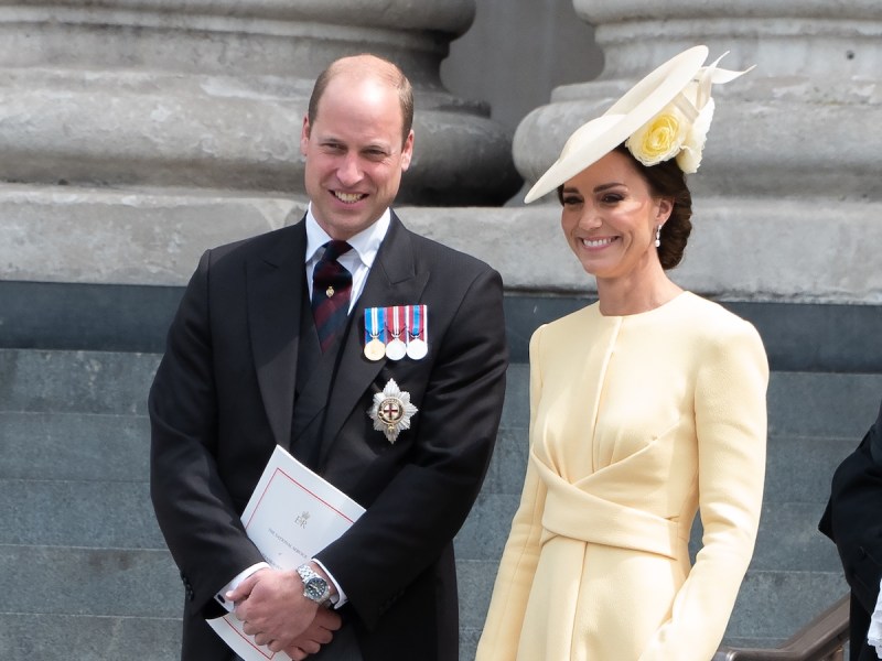 Prince William smiling in a black suit with Kate Middleton in a yellow dress and hat