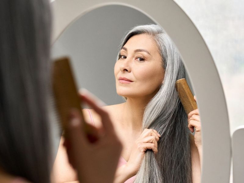 a woman looks in the mirror and brushes her long straight gray hair
