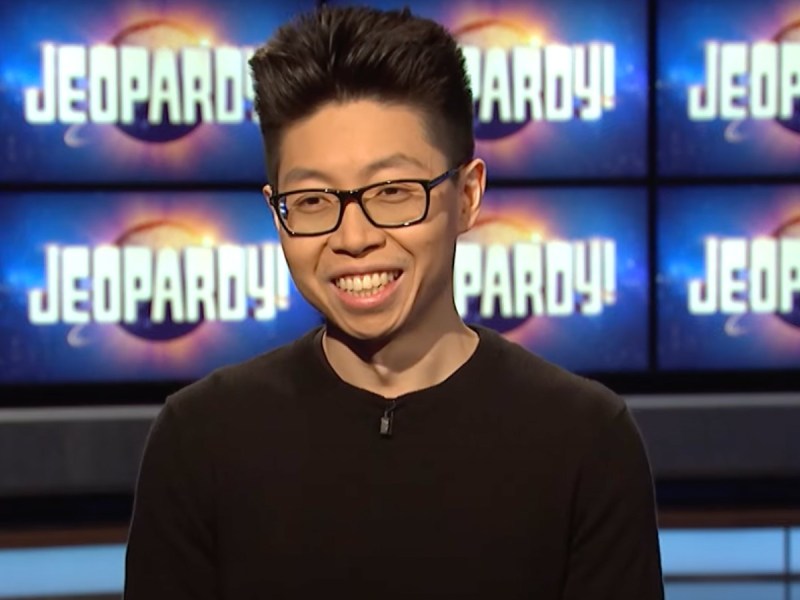 Andrew He smiles in black top with Jeopardy! screen in backdrop