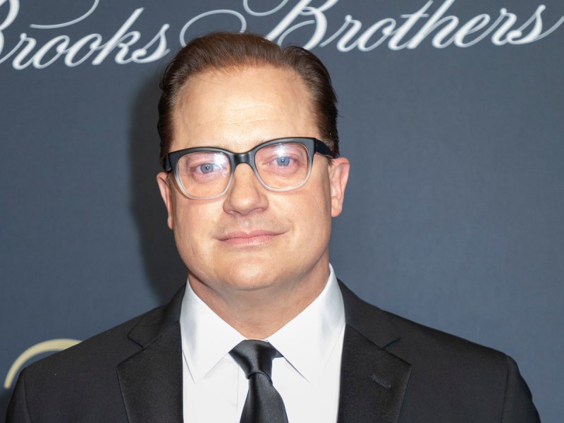 2018 photo of Brendan Fraser smiling in a black suit and tie with glasses on
