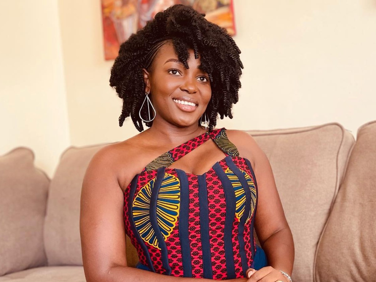 Charlot Magayi smiles while sitting on tan couch. She is wearing a red and blue striped dress with yellow accents
