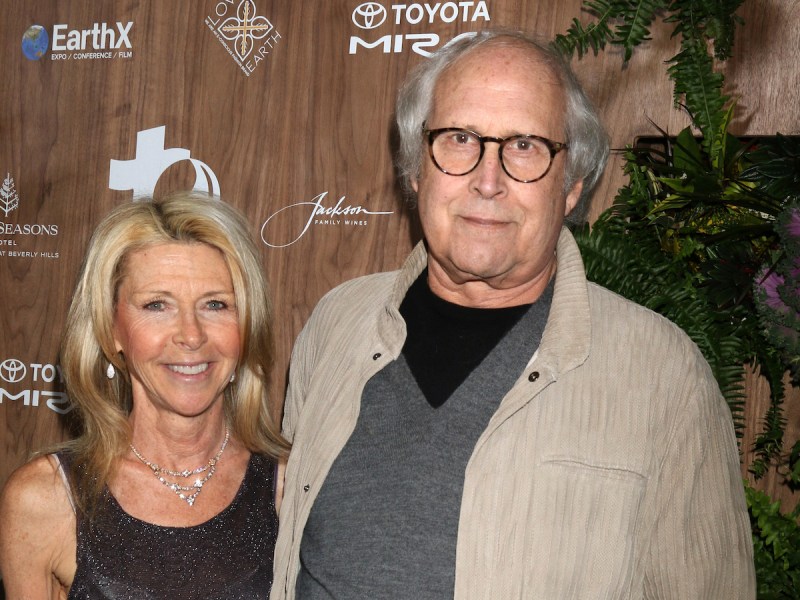 2019 photo of Chevy Chase in a grey shirt and tan coat with arm around Jayni Chase in a black dress