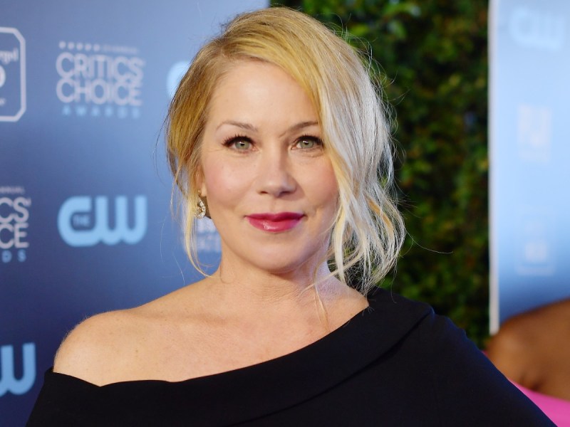 Christina Applegate smiles in off-shoulder black dress against blue backdrop