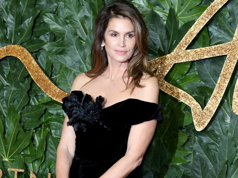 Cindy Crawford poses in black off-shoulder gown against green and gold backdrop