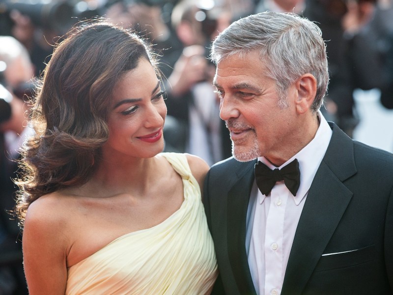 George Clooney (R) smiling in suit looking at Amal Clooney, who is wearing a one-shoulder cream colored/pale yellow gown
