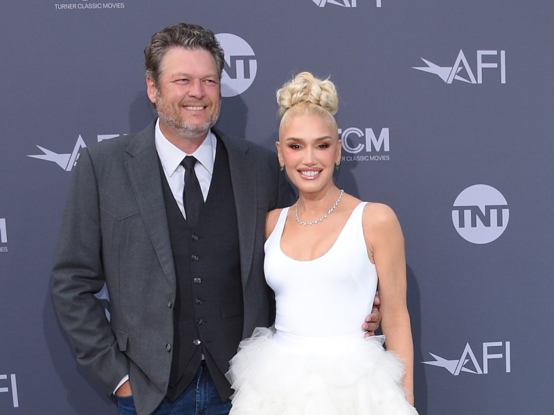 Blake Shelton in a grey suit smiling with arm around Gwen Stefani in a white dress
