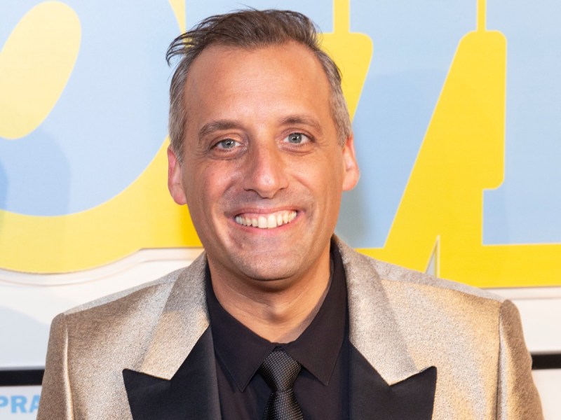 Joe Gatto smiling in black/gold suit and tie against blue and yellow backdrop