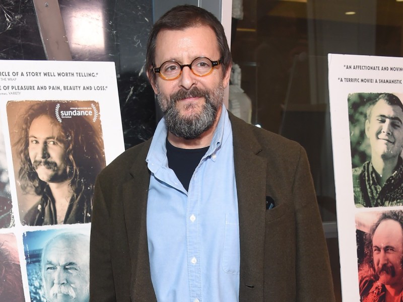 Judd Nelson smiles in blue button-down top and brown jacket