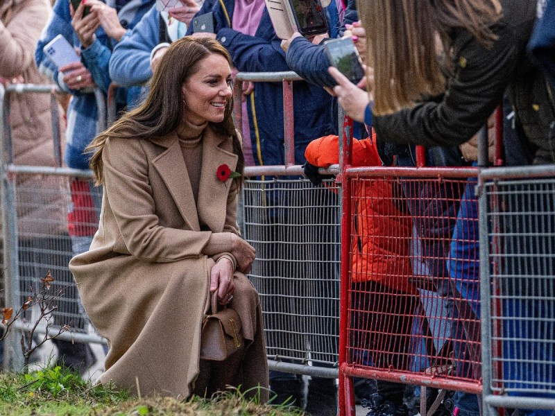 Kate Middleton kneels in tan coat to speak to child who is standing behind a fence