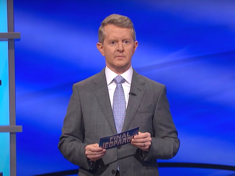 Ken Jennings in a grey suit holding a Final Jeopardy card