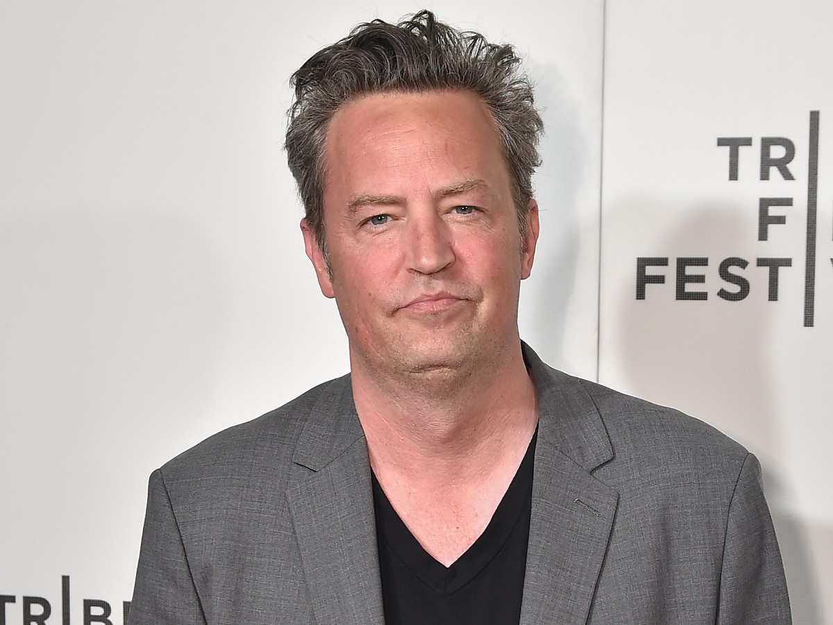 Matthew Perry smiles in gray suit jacket over black top against white backdrop