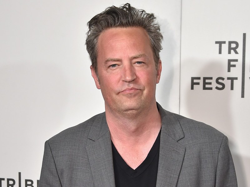 Matthew Perry smiles in gray suit jacket over black top against white backdrop