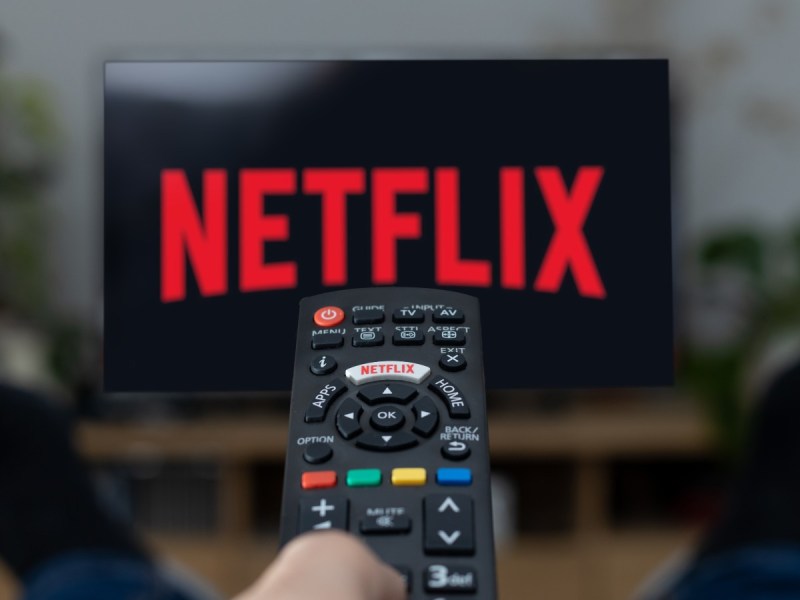 POV image of a hand pressing a remote control, which is pointed at a television with the Netflix logo on it