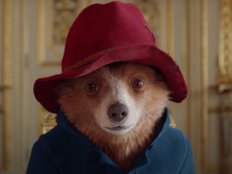 Paddington Bear sits at Buckingham Palace while wearing a red hat