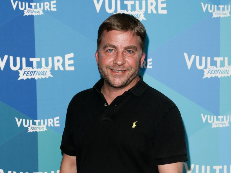 Peter Billingsley smiling in a black polo against a blue background