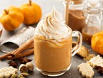 A pumpkin spice latte sits on a table surrounded by small pumpkins and cinnamon sticks
