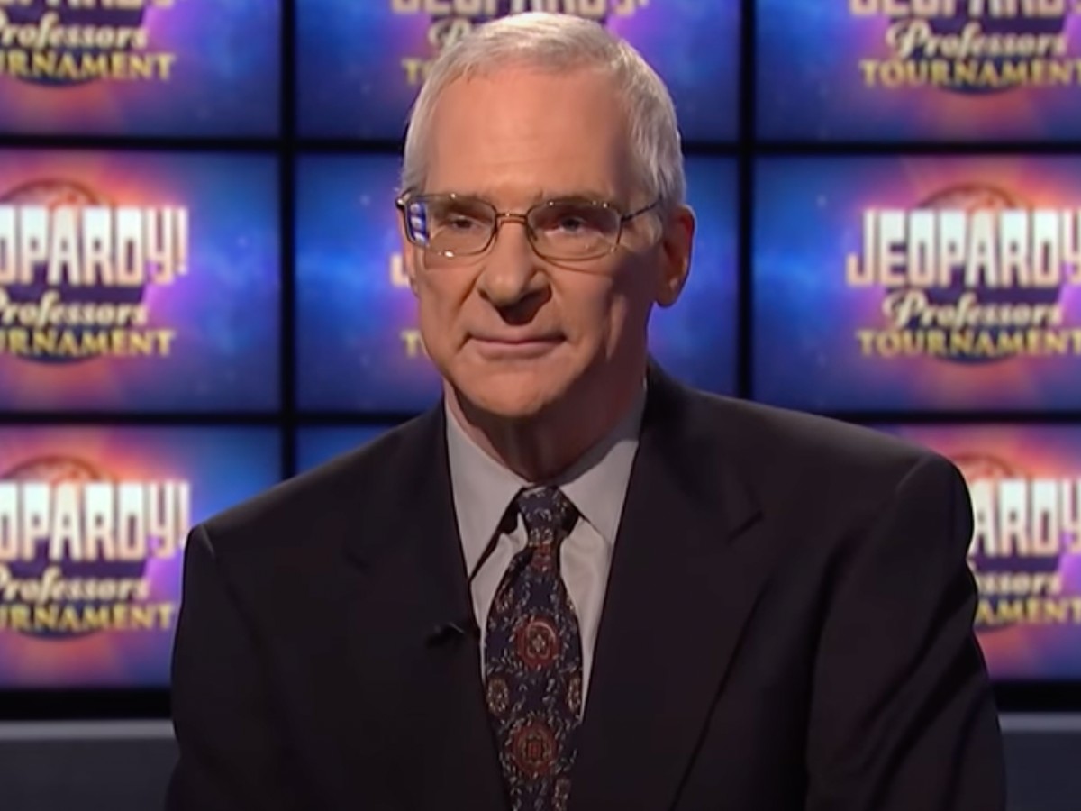 Sam Buttrey smirking in suit and tie against "Jeopardy!" screen background