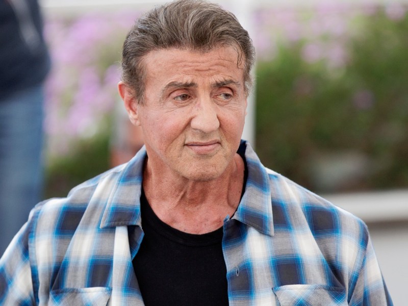 Sylvester Stallone posing in black shirt with a blue plaid overshirt