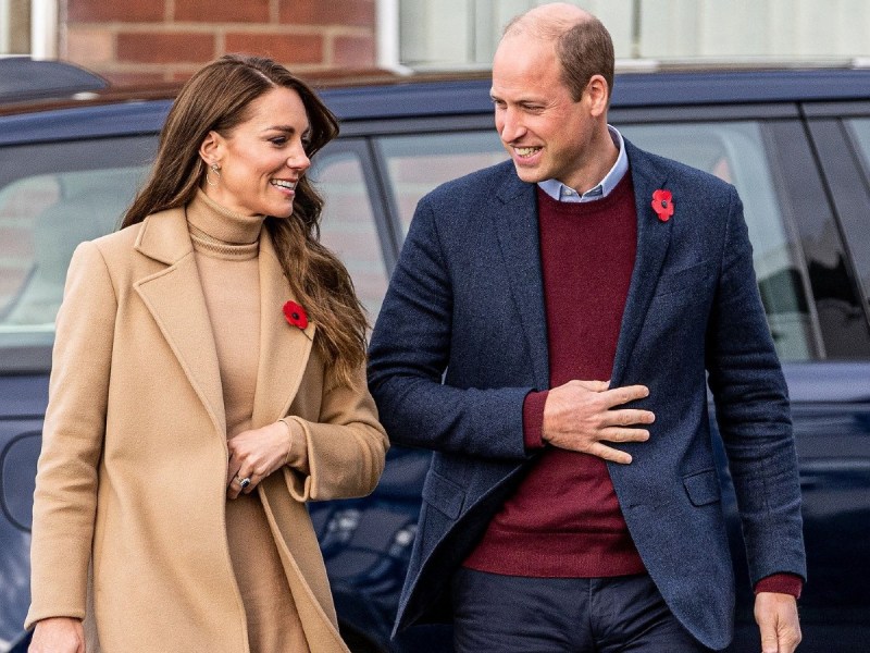 Kate Middleton (L) in tan coat with poppy pinned to lapel linking arms with Prince William, who is wearing a navy blue jacket over a red sweater