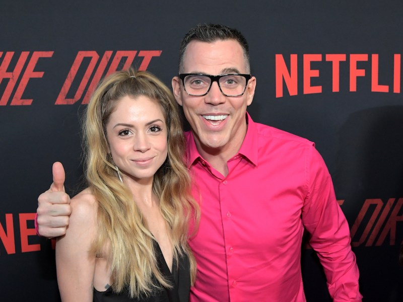 2019 photo of Steve-O in a pink shirt giving a thumbs up with arm around Lux Wright in a black dress