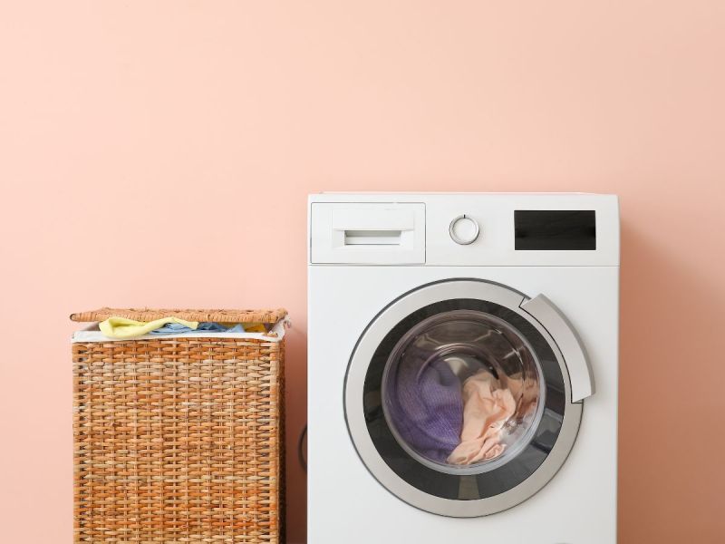 Washing machine with clothes inside next to hamper against peach-colored wall