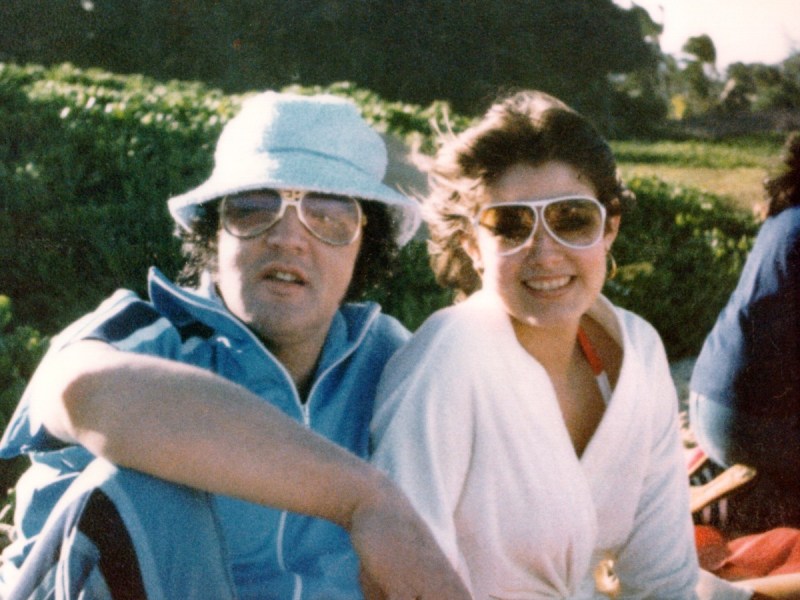 Elvis Presley (L) in blue top and white bucket hat sitting outside with Ginger Alden, who is wearing a pink top. Both are wearing sunglasses