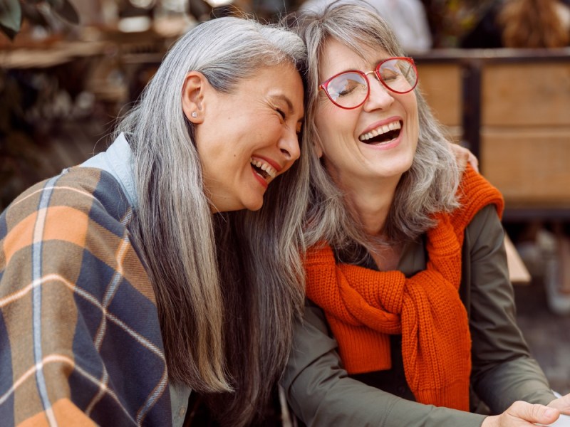 Two women laugh outdoors in fall clothing. Both women have gray hair