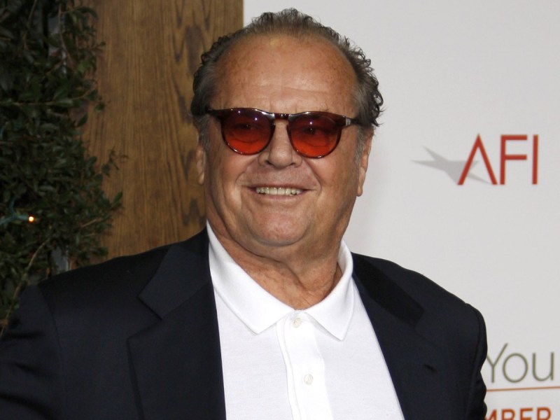 Jack Nicholson smiles in white top and black jacket with red-tinted sunglasses