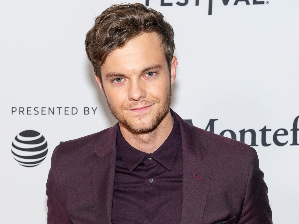 Jack Quaid smiles in burgundy suit set against white backdrop