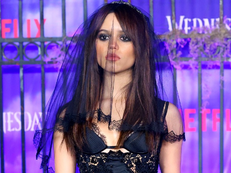 Jenna Ortega poses in black dress and black veil against purple backdrop
