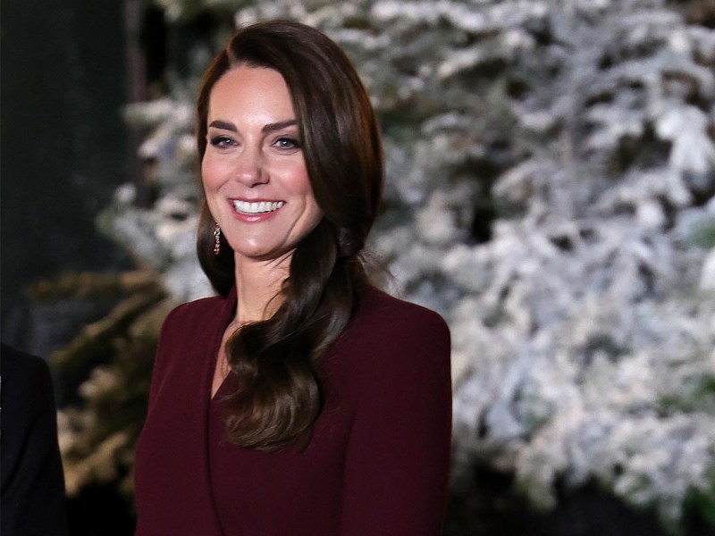 Kate Middleton smiling in a purple overcoat in front of a frosted pine tree.