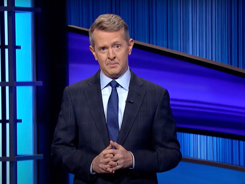 screenshot of Ken Jennings introducing Jeopardy in a navy suit and tie with an eyebrow raised and his hands clasped in front of him