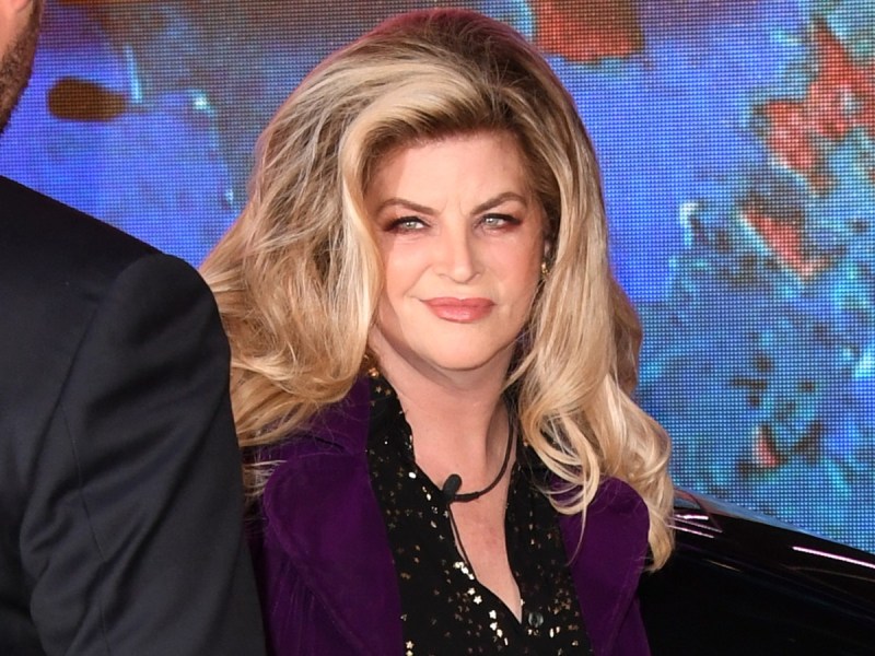 Kirstie Alley smiles in black top with purple sweater against blue backdrop
