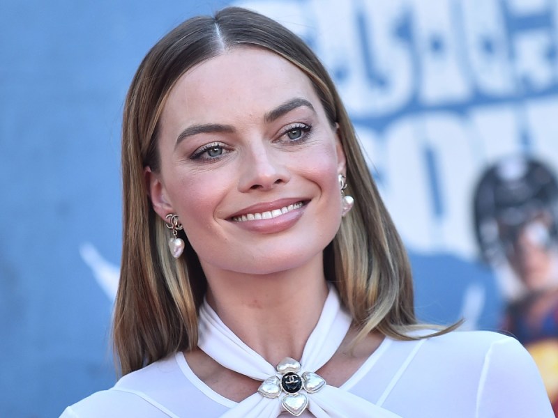 Closeup of Margot Robbie smiling in white top against blue backdrop