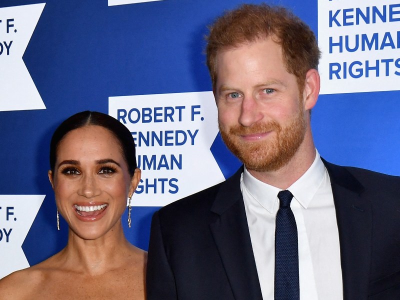 Prince Harry (R) and Meghan Markle smile against royal blue backdrop
