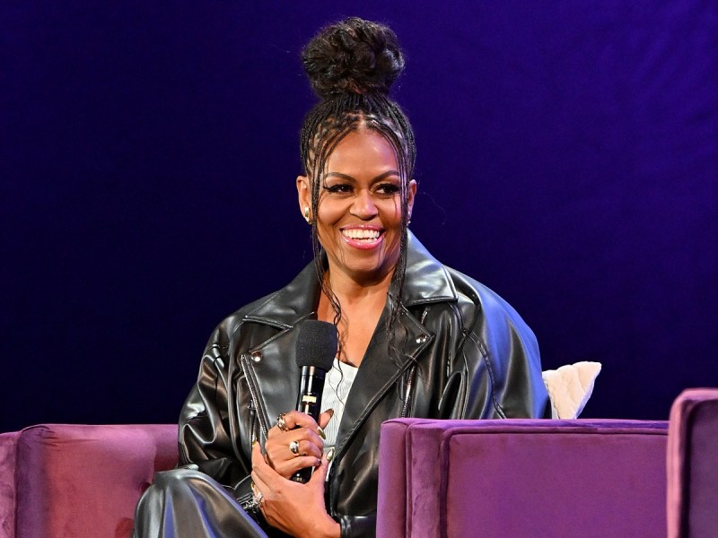 Michelle Obama smiles sitting in chair wearing black leather jacket