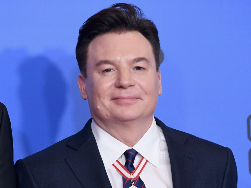 Mike Myers smiles in dark suit and tie against blue backdrop