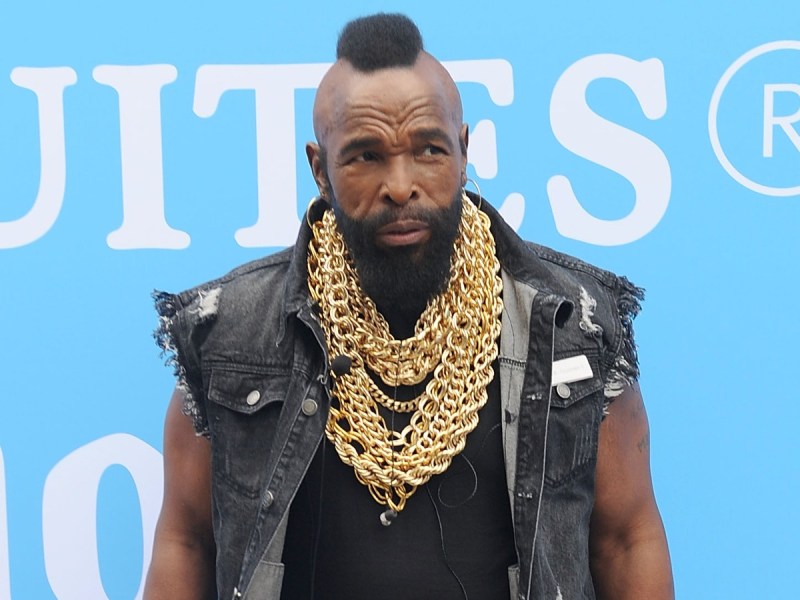 Mr. T looks off to the side wearing black top and gray denim vest against blue backdrop